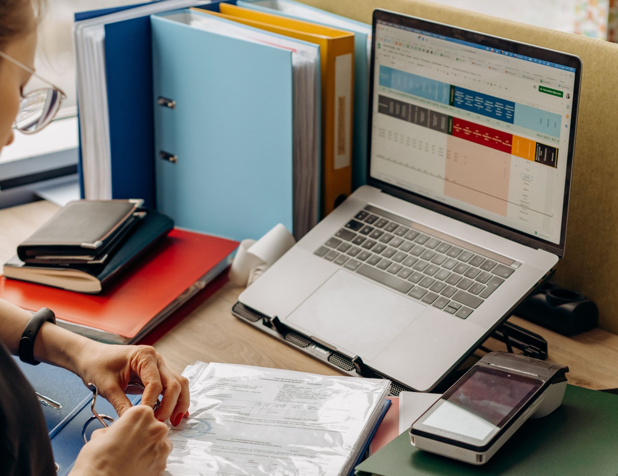 Person organizing papers at a desk with a laptop displaying a spreadsheet, several ring binders, notebooks, and a smartphone.